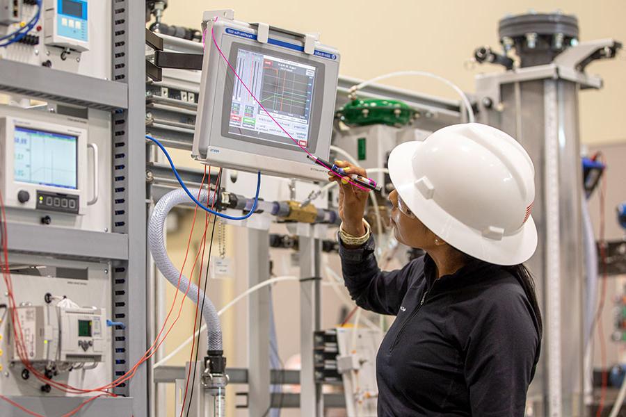 A person in a white hard hat looks at a monitor with various machines in the background